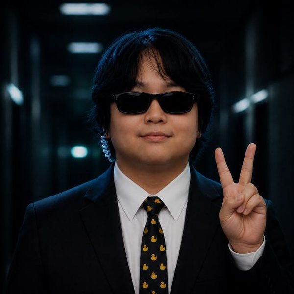Gen in a black suit, dark glasses and a rubber-duck tie, flashing a peace sign in a dim corridor.